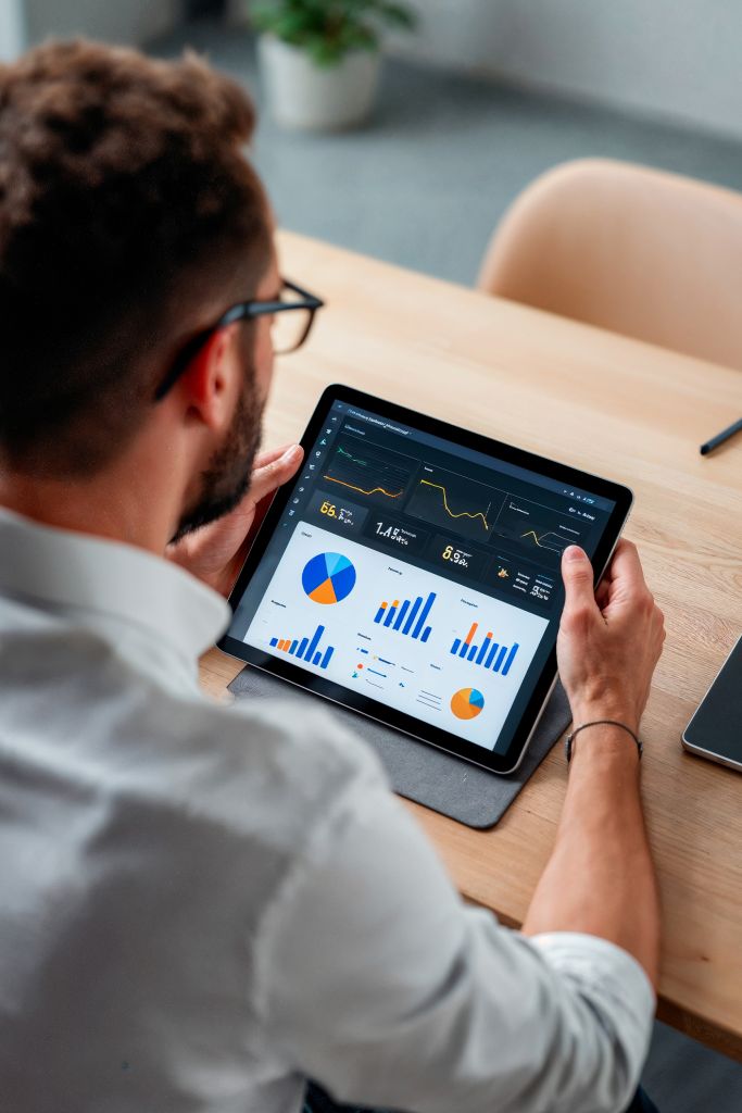Un homme portant des lunettes examine des graphiques et des diagrammes financiers, notamment des diagrammes à secteurs, des graphiques à barres et des graphiques linéaires, sur une tablette à son bureau tout en gérant Gestion des Réseaux Sociaux pour son entreprise.